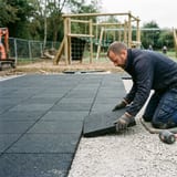 An installer laying a black rubber tile onto a prepared gravel base on a playground